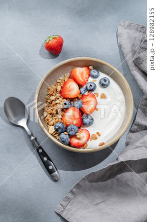 Natural yogurt with granola, blueberry and strawberry in a bowl on a blue background with spoon 128982155