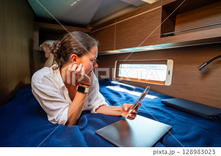 Young woman working with laptop and smartphone in the cabin of a sailing yacht near Croatia. Remote work, digital freedom and opportunity to live and work at sea. Young woman working with laptop and smartphone in the cabin of a sailing yacht near Croatia. Remote work, digital freedom and opportunity to live and work at sea. 128983023