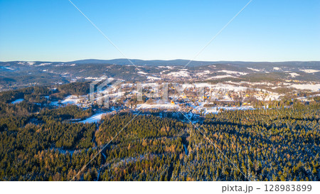 Panoramic view of Nova Ves nad Nisou showcasing the snow-covered landscape of the Jizera Mountains during a clear winter afternoon. Forests blanket the hills under a bright blue sky. 128983899
