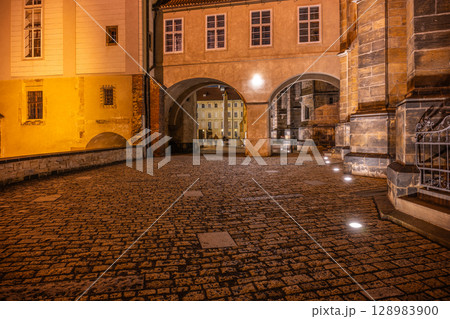 The covered bridge at Saint Vitus Cathedral in Prague Castle glows softly under lamp light, creating a magical atmosphere at night. Visitors explore the cobblestone paths nearby. 128983900