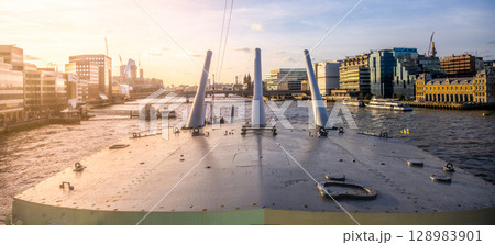 Turrets of HMS Belfast stand prominently along the River Thames, with the London skyline visible in the background during a peaceful sunset. 128983901