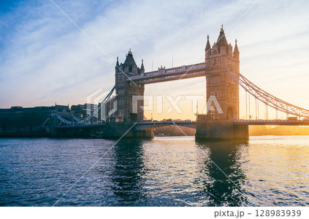 Tower Bridge stands majestically at sunrise, with warm hues illuminating its structure. The calm waters reflect the vibrant colors, showcasing a tranquil moment in the heart of London. 128983939