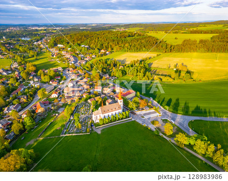 Bohutin Town is captured from above, revealing its charming layout nestled in lush greenery. Rolling hills and farmlands surround the peaceful village, emphasizing the natural beauty. Bohutin Town is captured from above, revealing its charming layout nestled in lush greenery. Rolling hills and farmlands surround the peaceful village, emphasizing the natural beauty. 128983986