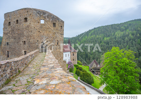 Visitors walk along the ancient stone bridge of Velhartice Castle in Czechia, surrounded by lush greenery and historical architecture on a cloudy day, capturing the essence of this scenic location. 128983988