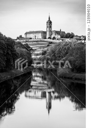 Chateau Melnik stands majestically on a hilltop, overlooking a tranquil river. The historic architecture is perfectly mirrored in the still water, showcasing its beauty in black and white. Chateau Melnik stands majestically on a hilltop, overlooking a tranquil river. The historic architecture is perfectly mirrored in the still water, showcasing its beauty in black and white. 128984010