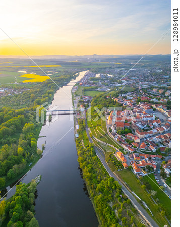 An aerial view captures Melnik City with its historic chateau perched above the serene Labe River, surrounded by green landscapes and rolling hills during golden hour. An aerial view captures Melnik City with its historic chateau perched above the serene Labe River, surrounded by green landscapes and rolling hills during golden hour. 128984011