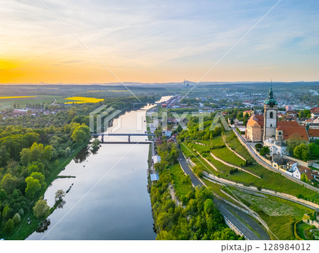 Aerial view captures Melnik city and its historic chateau perched above the tranquil Labe River. The golden hour illuminates the landscape, showcasing lush greenery and flowing waters. Aerial view captures Melnik city and its historic chateau perched above the tranquil Labe River. The golden hour illuminates the landscape, showcasing lush greenery and flowing waters. 128984012