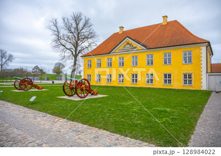 The vibrant yellow Commander's House stands proudly in Kastellet, Copenhagen, surrounded by historical cannons. Visitors enjoy the serene green space with a cloudy sky above. 128984022
