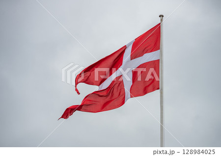 The Denmark flag flies proudly on a pole, fluttering in the wind under a cloudy sky. This vibrant red and white design represents the national identity and culture of Denmark. The Denmark flag flies proudly on a pole, fluttering in the wind under a cloudy sky. This vibrant red and white design represents the national identity and culture of Denmark. 128984025