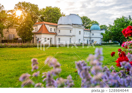 Visitors explore the Stefanik Observatory on Petrin Hill in Prague. The charming building is surrounded by blooming flowers and greenery, creating a serene atmosphere during sunset. 128984044