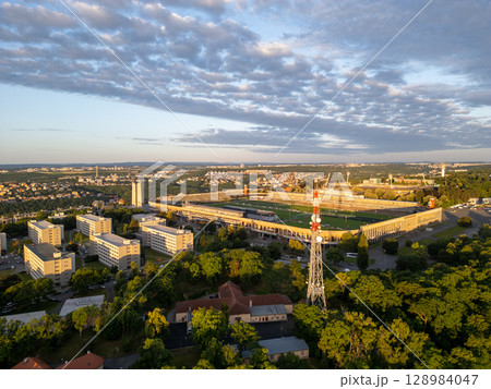Great Strahov Stadium, surrounded by lush greenery, showcases its vastness during sunset in Prague. Buildings and trees create a beautiful landscape, highlighting the stadium's architectural grandeur. Great Strahov Stadium, surrounded by lush greenery, showcases its vastness during sunset in Prague. Buildings and trees create a beautiful landscape, highlighting the stadium's architectural grandeur. 128984047
