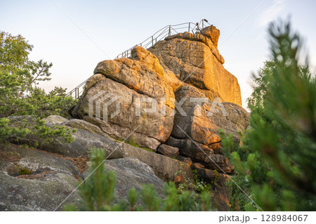 The sun sets over Popov Cliffs, casting a warm glow on the sandstone rock formations. Lush greenery surrounds the cliffs, creating a tranquil evening atmosphere in Czechia. 128984067