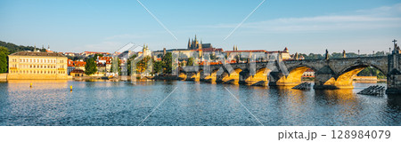Breathtaking panorama showcases Prague Castle rising above the Vltava River, with the iconic Charles Bridge spanning the calm waters, reflecting the morning light in a serene cityscape. 128984079