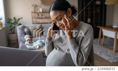 Happy pregnant black mom working remotely on laptop. Online video call. Engrossed in her video call, the joyful mom balances her laptop and baby bump with ease. 128986633