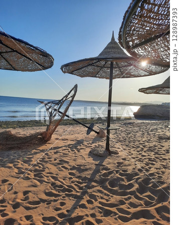 Sandy Beach with Woven Parasols and Calm Blue Ocean at Sunset Sandy Beach with Woven Parasols and Calm Blue Ocean at Sunset 128987393