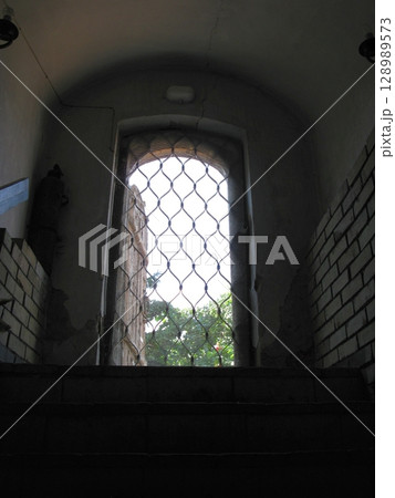 A narrow arched window with a metal grid in a brick walled cave, allowing a glimpse of greenery outside under a dimly lit interior 128989573