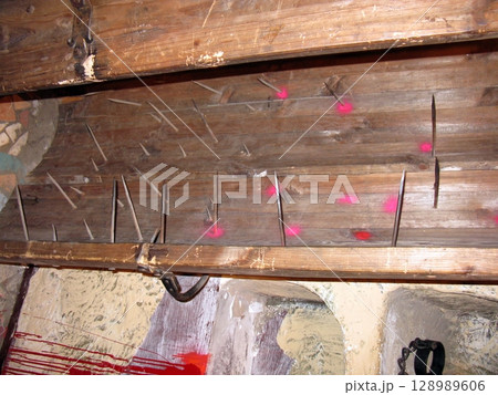 A wooden ceiling panel with protruding spikes and red stains, mounted in a stone walled room, evoking a chilling historical torture trap A wooden ceiling panel with protruding spikes and red stains, mounted in a stone walled room, evoking a chilling historical torture trap 128989606