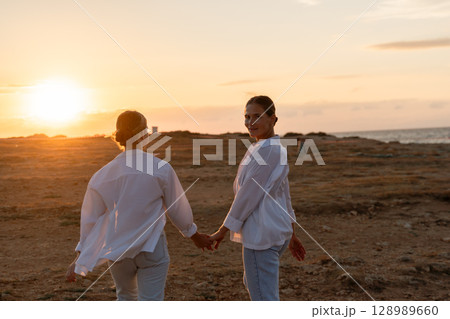 Couple Sunset Beach Walk - Two people in white clothing walk hand-in-hand towards a sunset on a beach. 128989660