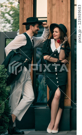 Man tipping hat and smiling at woman in vintage black dress standing by wooden wall Man tipping hat and smiling at woman in vintage black dress standing by wooden wall 128989683