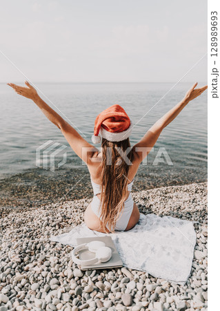 A woman wearing a red Santa hat is sitting on a towel on a beach. She is holding her arms up in the air, as if she is celebrating something. A woman wearing a red Santa hat is sitting on a towel on a beach. She is holding her arms up in the air, as if she is celebrating something. 128989693