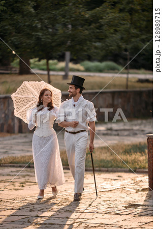 Man and woman in white vintage clothes walking with lace umbrella in park 128989715