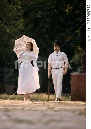 Woman walking ahead with parasol while man in top hat follows behind 128989717