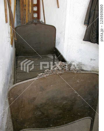 A rustic metal bed frame with ornate headboards tucked in a narrow, whitewashed room alongside tools and a hanging coat A rustic metal bed frame with ornate headboards tucked in a narrow, whitewashed room alongside tools and a hanging coat 128989860