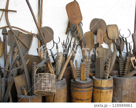 A collection of weathered wooden tools and implements arranged against a white wall with rustic barrels and a woven basket A collection of weathered wooden tools and implements arranged against a white wall with rustic barrels and a woven basket 128989861