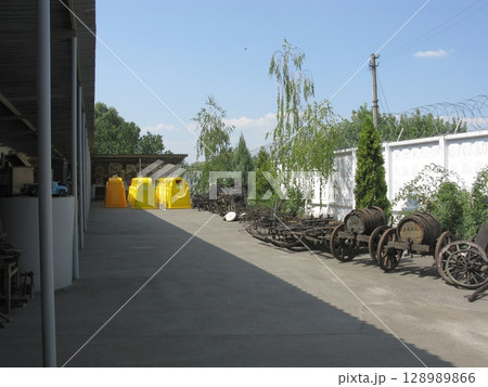 An outdoor display of weathered wooden carts and barrels near a white fence with trees under a clear blue sky 128989866