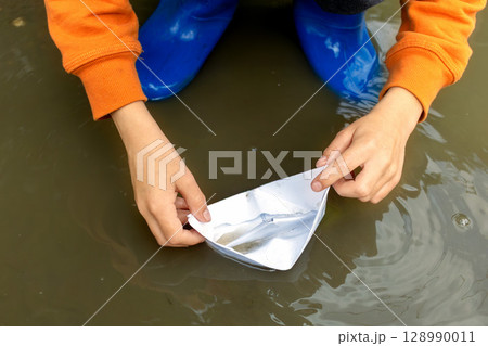 Child in blue rain boots gently sets paper boat afloat in puddle. Children playing outdoors. Close-up 128990011
