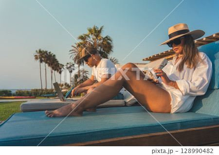 A woman wearing a straw hat and sunglasses is sitting on a blue lounge chair, remote work concept A woman wearing a straw hat and sunglasses is sitting on a blue lounge chair, remote work concept 128990482