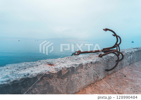A rusty anchor is sitting on a concrete wall next to the ocean 128990484