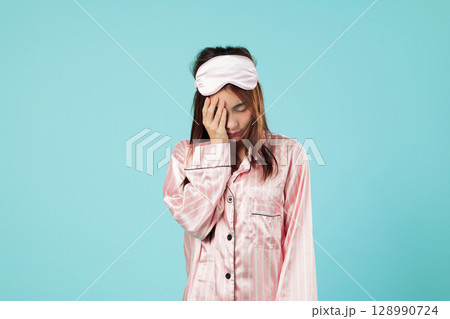Tired asian woman sleep eye yawning covering in pajamas with hand on her face, eyes closed, waking up gloomy looking tired after a party covers her face with her hand in pain, isolated blue background 128990724