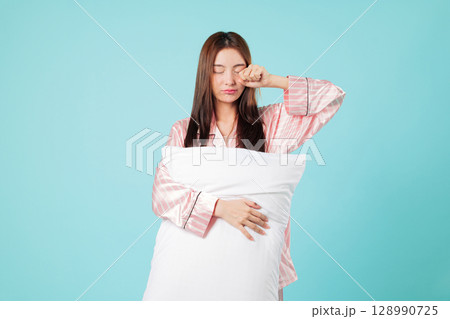 Asian woman yawns, rubbing eye after restful sleep, she is waking up, holding her pillow in good mood after a sound sleep, headshots isolated studio blue background Asian woman yawns, rubbing eye after restful sleep, she is waking up, holding her pillow in good mood after a sound sleep, headshots isolated studio blue background 128990725