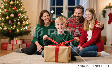 Happy boy opening Christmas gift surrounded by smiling family in festive living room with decorated tree and wrapped presents 128990831