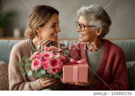 Happy mother and daughter joyfully share a special moment, exchanging a beautiful flower bouquet and a thoughtful gift. 128992158