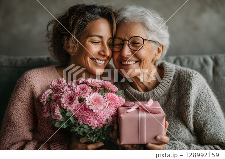 A loving mother and her happy daughter share a warm embrace, holding a vibrant pink flower bouquet and a thoughtful gift. 128992159