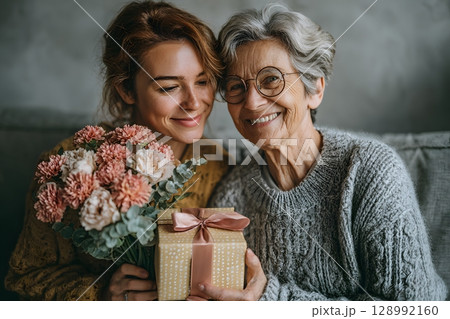 Joyful mother and daughter share a special day, smiling brightly while holding a beautiful flower bouquet and a thoughtful gift. 128992160