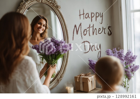A mother holds purple flowers admiring her reflection, celebrating Mother's Day, as a child watches nearby. 128992161