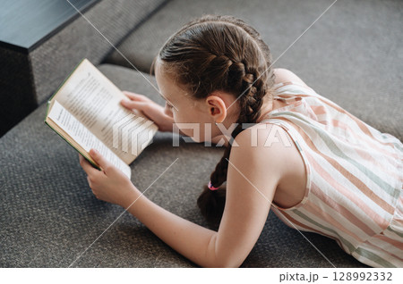 A girl comfortably lying on a sofa, engrossed in reading a book, depicting relaxation and quiet leisure A girl comfortably lying on a sofa, engrossed in reading a book, depicting relaxation and quiet leisure 128992332