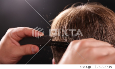 Young Man with Beard at Barber Shop. Barber is Preparing his Haircut. Dark Atmosphere, Black Background. Concept of Barber and a Client. Young Man with Beard at Barber Shop. Barber is Preparing his Haircut. Dark Atmosphere, Black Background. Concept of Barber and a Client. 128992739