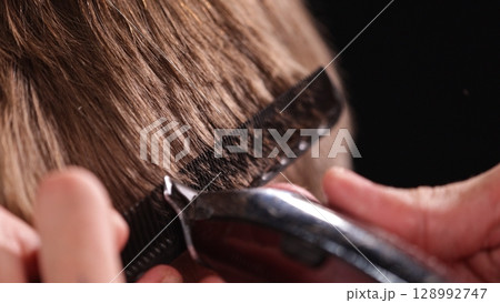 Young Man with Beard at Barber Shop. Barber is Preparing his Haircut. Dark Atmosphere, Black Background. Concept of Barber and a Client. Young Man with Beard at Barber Shop. Barber is Preparing his Haircut. Dark Atmosphere, Black Background. Concept of Barber and a Client. 128992747