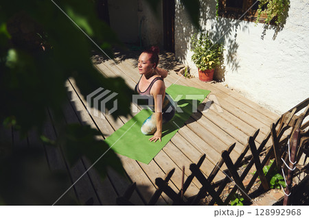 Woman Performing Yoga on Outdoor Deck Surrounded by Natural Light and Greenery 128992968
