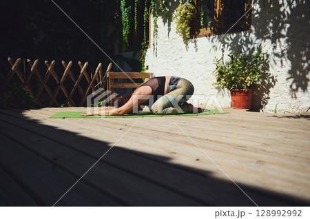Woman Practicing Yoga in Sun-Lit Outdoor Deck Surrounded by Foliage 128992992