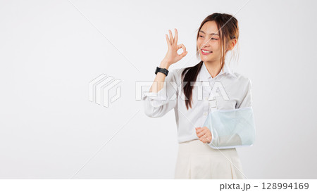 Happy young asian woman injury arm broken with a soft splint and sling makes an ok gesture after her accident showing she is doing fine and recovering well from her accident, isolated white background 128994169