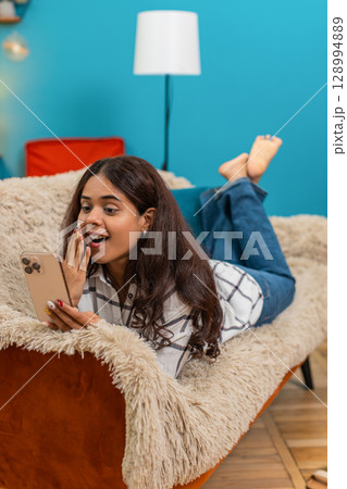 Indian woman lying with smartphone celebrates good news message raises fist excited joyful indoors 128994889