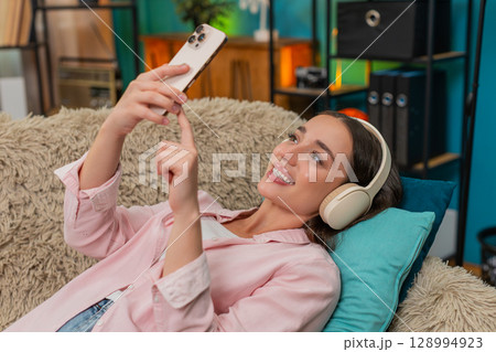 Young woman scrolling social media on smartphone with wireless headphones enjoying restful downtime 128994923