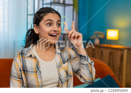 Indian woman sitting on sofa raising finger sudden solution Eureka expression discovering idea happy 128995004