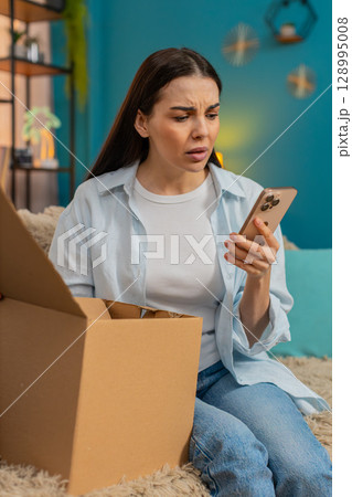 Young woman sitting on sofa unpacking delivery finding wrong item disappointed shaking head sadly 128995008
