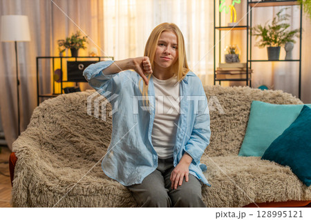 Sad young woman thumbs down at home, displeased face, negativity, critical mood, rejecting sign Sad young woman thumbs down at home, displeased face, negativity, critical mood, rejecting sign 128995121